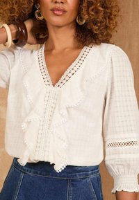 Blusa bianca con volant, dettagli in pizzo e scollatura a V. Presenta maniche a tre quarti e un tessuto texturizzato. Abbinata con una gonna di jeans.