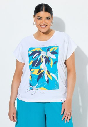 Smilende kvinde med mørkt hår iført hvid t-shirt med blå og gul blomsterprint og blå bukser, stående mod en hvid baggrund.