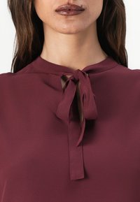 Mørkerød bluse med høj halsudskæring og detalje med forbinding. Stoffet ser glat ud med en subtil glans.
