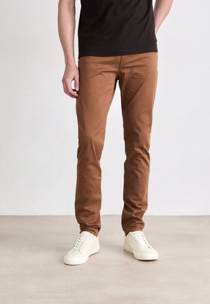 Chinos de algodón marrones con un corte ajustado, con una textura suave y bolsillos frontales, combinados con zapatillas de deporte en color blanco roto.