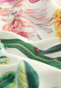 Floral bedrukte stof met levendige roze, oranje en groene tinten; zachte textuur met zichtbare plooien en een crème achtergrond.