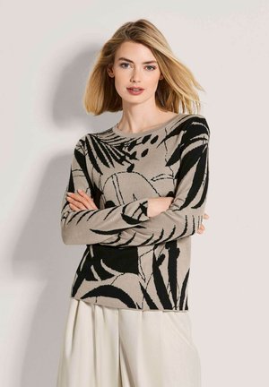 Frau mit blondem Haar, die einen beigefarbenen und schwarz gemusterten Pullover und weiße Hosen trägt, steht mit verschränkten Armen vor einem einfarbigen Hintergrund.