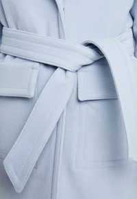 Manteau en laine bleu clair avec ceinture nouée et deux poches à rabat rectangulaires à la taille, montrant un gros plan de la zone médiane.