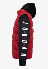 Rød puffer vest med sort hætte, der har en tekstureret overflade. Venstre ærme har "JORDAN" i store hvide bogstaver. Sorte ribbede manchetter.