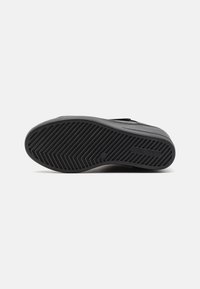 Suela de zapatilla negra con un patrón texturizado en zigzag y un logo. El material parece ser goma, con áreas lisas y estriadas para mayor tracción.