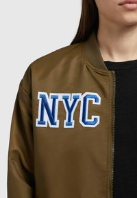 Oliivroheline bomber-jakk, millel on lukuga eesmine osa, soonikkoes krae ja sinise ja valge värviga brodeeritud "NYC" logo vasakul rinnal.