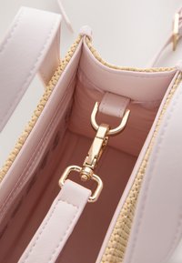 Nahaufnahme des hellrosa Innenraums einer Handtasche mit goldfarbenem Verschluss, der den abnehmbaren Riemen an der inneren Schlaufe der Tasche befestigt.