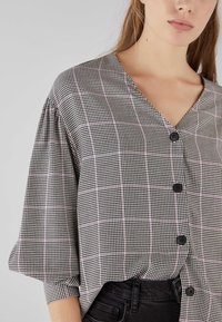 Blusa a scacchi bianco e nero con scollo a V, maniche corte a palloncino e quattro bottoni neri sul davanti. Progettata con una vestibilità ampia.
