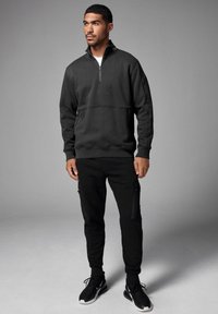 Felpa nera con zip e colletto alto, caratterizzata da una cucitura sul petto e tasche laterali; abbinata a pantaloni neri a gamba affusolata e scarpe da ginnastica.
