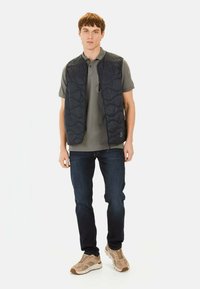 Gequilte marineblauwe vest met een rits aan de voorkant, gedragen over een grijze polo, in combinatie met donkere jeans en beige sneakers. Heeft een gladde textuur en subtiele patronen.