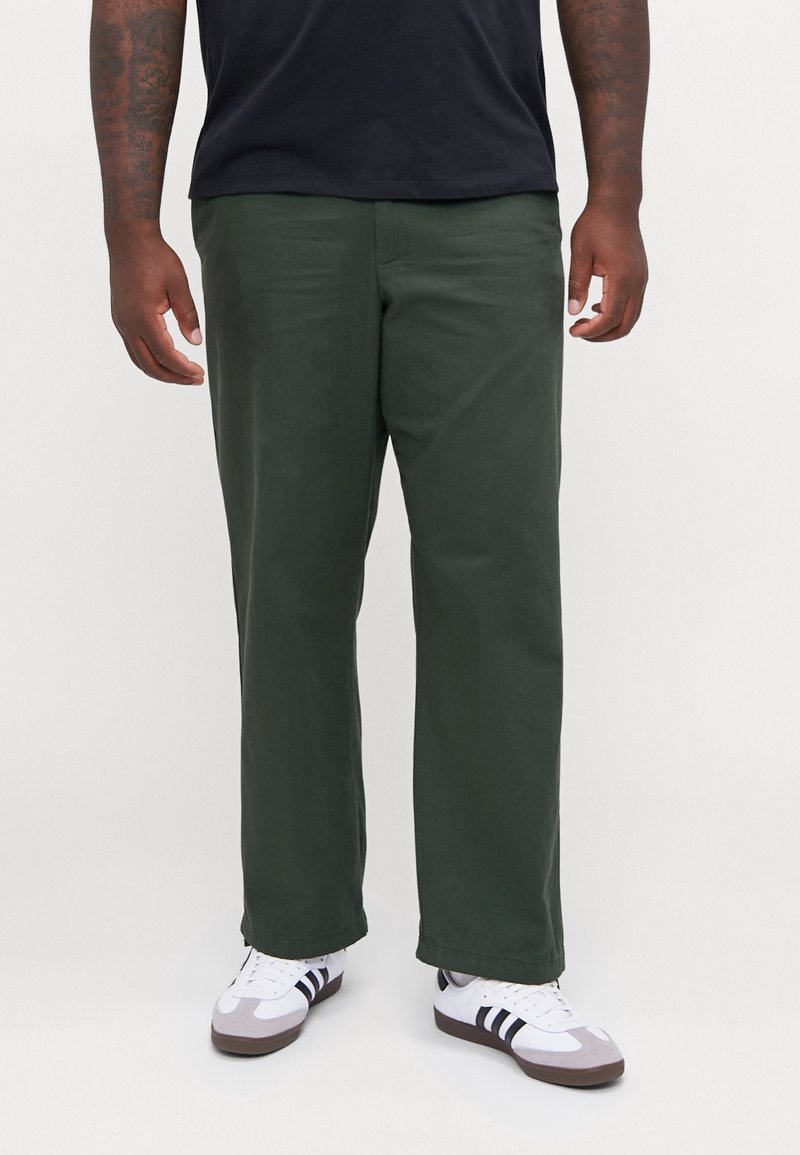 Dunkelgrüne, gerade geschnittene Hose aus einem weichen Stoff mit einer subtilen Textur. Kombiniert mit weißen Sneakers und einem schwarzen Hemd.