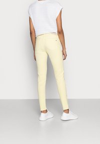 Jeans ajustados en denim amarillo claro, con un diseño clásico de cinco bolsillos y un pequeño detalle de marca en el bolsillo trasero. Llevados con zapatillas blancas.