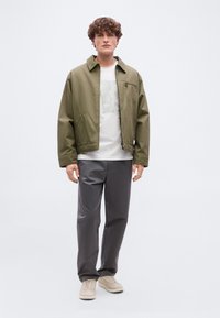 Νεαρός άνδρας με σγουρά μαλλιά που φοράει μπουφάν ελιάς, λευκό μπλουζάκι με γραφικό σχέδιο, γκρι παντελόνι και μπεζ αθλητικά παπούτσια, στέκεται πάνω σε απλό φόντο.