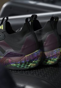 Paire de baskets noires sans lacets avec des semelles à motifs violet, vert néon et multicolore, placée sur une surface noire texturée.