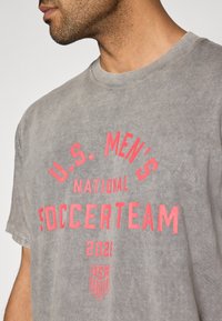 Šedé bavlněné tričko s červeným potiskem "U.S. MEN'S NATIONAL SOCCERTEAM 2020" a logem U.S. Soccer ve spodní části. Kulatý výstřih.