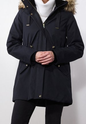 Personne portant un manteau d'hiver noir avec une capuche bordée de fourrure sur un pull à col roulé blanc, mains jointes devant.