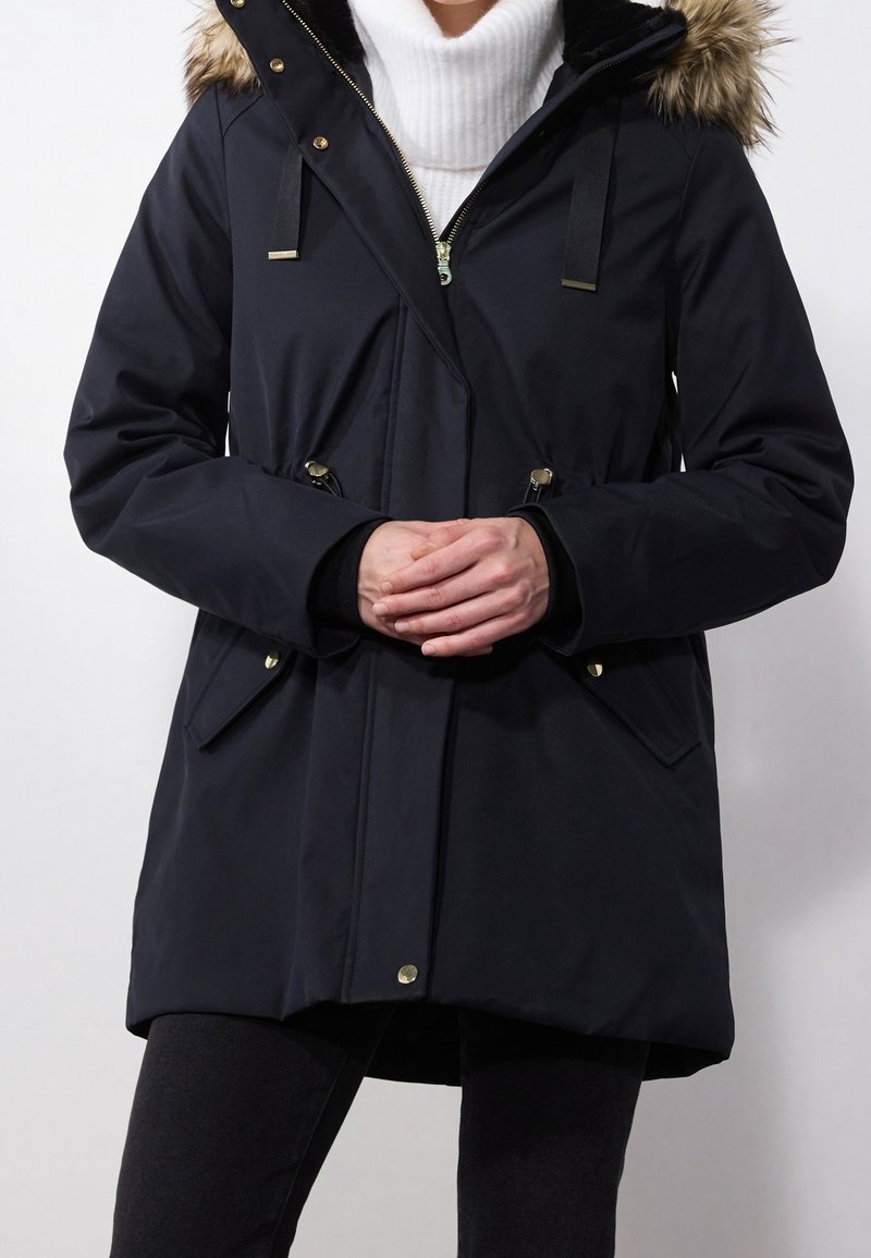 Personne portant un manteau d'hiver noir avec une capuche bordée de fourrure sur un pull à col roulé blanc, mains jointes devant.