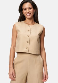 Top beige court sans manches avec un col rond, cinq boutons sur le devant, et une texture lisse, associé à un pantalon taille haute beige.