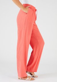 Pantalons larges corail en tissu texturé. Dotés d'une taille élastique avec cordon de serrage et de poches latérales. Associés à des sandales blanches.