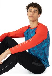 Langärmliges Sportoberteil mit einem blauen und türkisfarbenen Pixelmuster und roten Ärmeln, aus dehnbarem Stoff, kombiniert mit schwarzen Hosen.