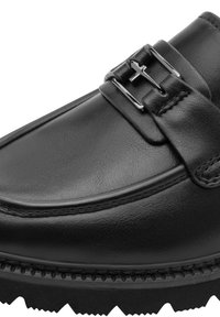 Mocassin en cuir noir avec une boucle métallique argentée et un détail en forme de croix sur le dessus, avec une couture soignée et une semelle robuste.