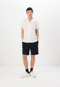 Maglietta bianca a maniche corte in maglia con un motivo testurizzato, abbinata a pantaloni corti blu navy e sneakers bianche. Collo semplice e vestibilità rilassata.