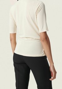 Světle béžový top s krátkým rukávem, s přiléhavým střihem, jemnou texturou a subtilními postranními větracími otvory, kombinovaný s černými dobře střiženými kalhotami.