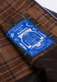 Καφέ καρό ύφασμα με κίτρινες λεπτομέρειες και μπλε ετικέτα που περιλαμβάνει λευκά σχέδια ενδυμάτων. Το κείμενο στην ετικέτα γράφει "Ο Paul Smith αγαπά τον Barbour."