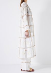 Lange weiße Tunika mit gestickten Blumenmustern, beigefarbenen Akzenten und offenem Vorderteil. Kombiniert mit weißen Hosen und braunen Sandalen.
