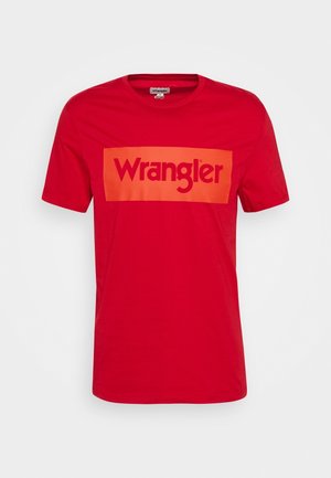 Red cotton t-shirt featuring a large orange rectangular graphic with the text "Wrangler" in bold, contrasting font. Short sleeves and crew neck.