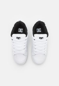 Hvite sneakers med en glatt læroverdel, svart stofffôr, flate hvite snøringer og en fremtredende DC-logo på tungen og siden.
