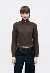 Καφέ κοντή ζακέτα με φερμουάρ και ψηλό γιακά, φτιαγμένη από μαλακό υφασμάτινο υλικό με υφή, σε συνδυασμό με σκουρόχρωμο τζιν υψηλής μέσης και μαύρη ζώνη.