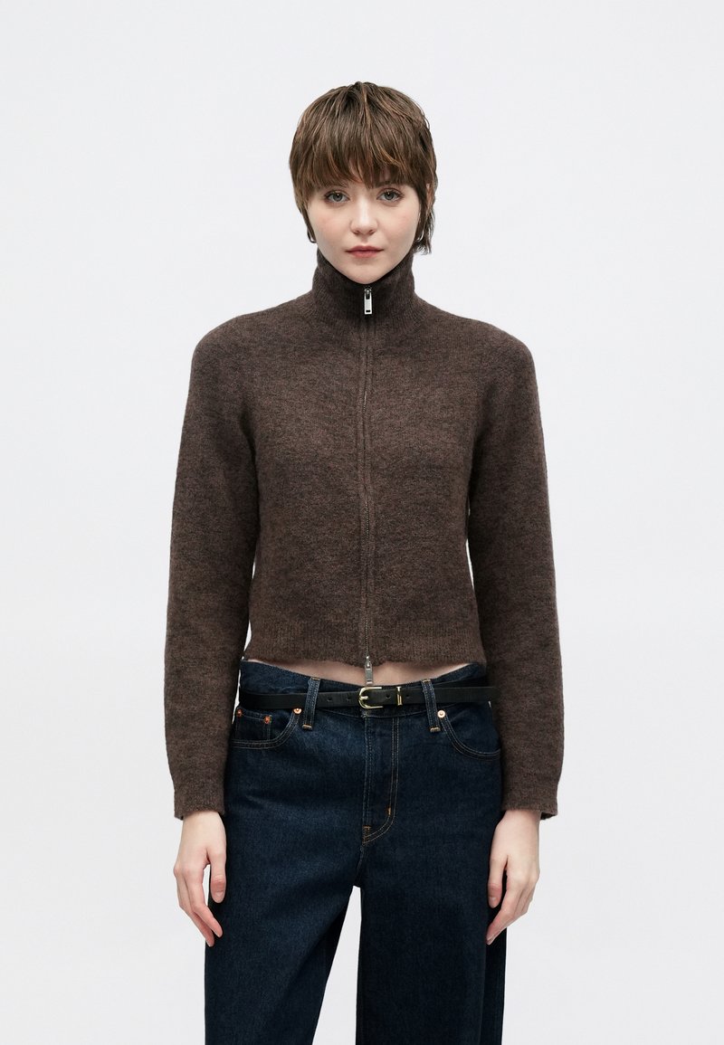 Καφέ κοντή ζακέτα με φερμουάρ και ψηλό γιακά, φτιαγμένη από μαλακό υφασμάτινο υλικό με υφή, σε συνδυασμό με σκουρόχρωμο τζιν υψηλής μέσης και μαύρη ζώνη.