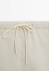 Pantalon beige avec une taille élastique et un cordon noué à l'avant, coutures visibles et poches latérales.