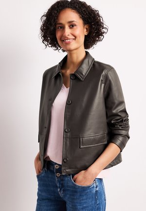 Femme souriante aux cheveux bouclés portant une veste en cuir gris foncé sur un haut rose clair, mains dans les poches d'un jean bleu, debout sur un fond blanc.