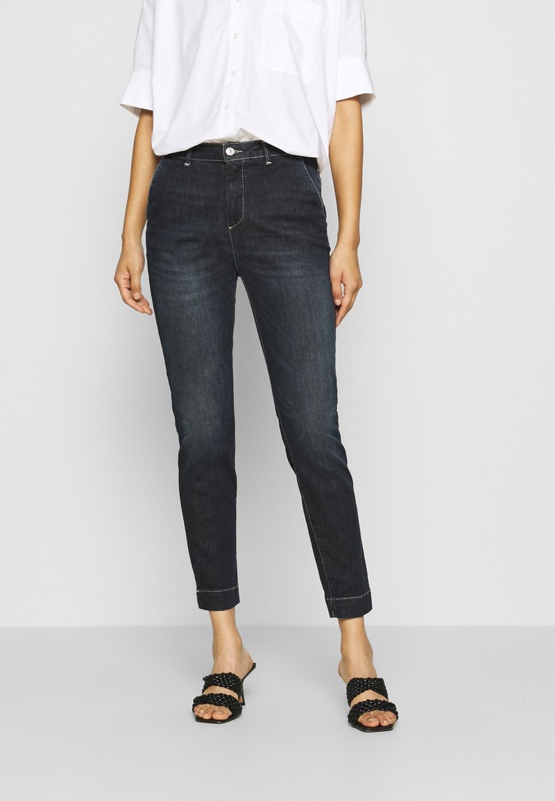 Tummansiniset farkut, joissa on kapea malli ja nilkkapituisuus, klassinen viiden taskun malli ja kontrastiväriset ompelupisteet. Yhdistetty valkoisen nappipaidan ja mustien liukukenkien kanssa.