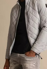 Giacca trapuntata grigio chiaro con chiusura a zip, tasche laterali e un patch accentato nero. Indossata sopra una maglietta nera e jeans grigi strappati.