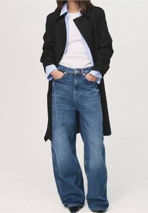 Person iført vide blå jeans, hvid T-shirt, blå skjorte med knapper og sort lang frakke med hænderne i lommerne mod en ensfarvet baggrund.