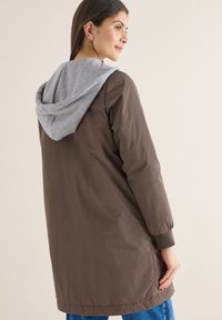 Femme portant une longue veste marron avec une capuche grise, regardant sur le côté, la main posée sur le côté de la veste.