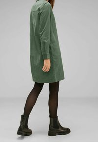 Groene shirtjurk van gestructureerde stof. Heeft lange mouwen, een kraag en zijzakken. Gepaard met zwarte enkellaarsjes en panty's.