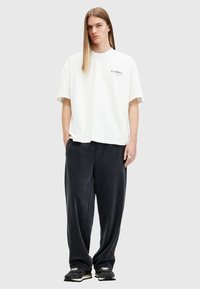White oversized t-shirt with logo on chest, paired with dark grey, soft, wide-leg pants. Model wears black sneakers.