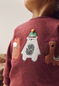 Pull bordeaux avec des motifs d'animaux brodés : un renard portant une écharpe à carreaux, un ours avec un chapeau vert, et un chien marron avec des détails texturés.