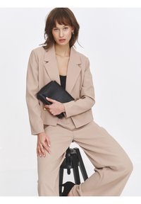 Beige tailored blazer and wide-leg trousers, made of soft fabric, paired with a black clutch featuring gold hardware accents.