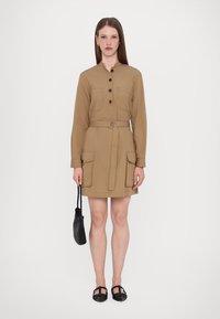 Abito camicia color khaki con maniche lunghe, chiusura a bottoni davanti, due tasche anteriori, vita svasata e una cintura. Abbinato a una piccola borsa nera.
