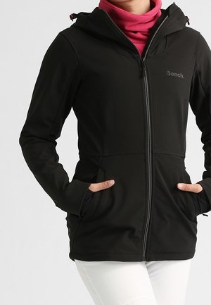 Osoba ubrana w czarną kurtkę z kapturem zapinaną na zamek, ręce w kieszeniach, pod nią różowy golf i białe spodnie, na jednolitym tle.