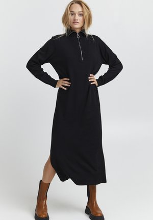 Black, long-sleeve dress with a zippered high collar, made from soft fabric; features side slits and falls below the knees.