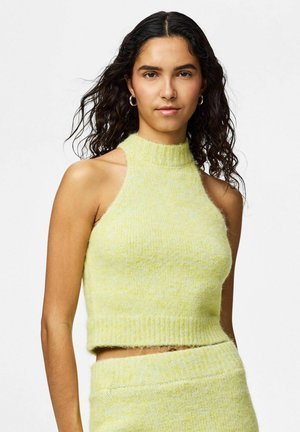 Donna con capelli ricci scuri che indossa un top corto senza maniche in maglia giallo pallido e una gonna abbinata, rivolta in avanti su uno sfondo bianco.