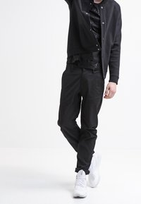 Černá bomber bunda s knoflíky, vrstvená přes černou vzorovanou košili, doplněná černými kalhotami a bílými teniskami. Hladká tkanina a volný střih.