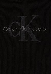 Testo grigio in rilievo "Calvin Klein Jeans" e "CK" in un carattere stilizzato su un tessuto nero a coste. Texture liscia con una leggera lucentezza.