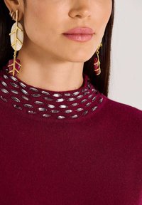 Frau mit langen goldenen und roten blattförmigen Ohrringen und einem burgunderfarbenen Pullover mit glänzenden ovalen Verzierungen am hohen Ausschnitt.